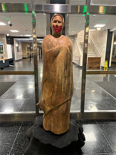 Carved wooden sculpture of a cloaked human figure with a painted face and red lips displayed in a building lobby.